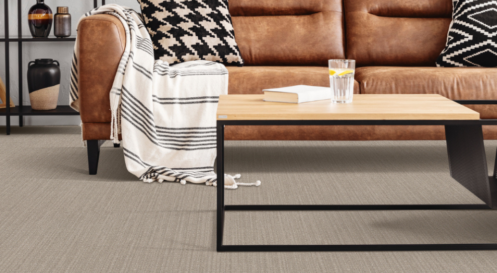 A modern living room with grey carpet floors, a brown leather sofa, patterned throw pillows, a light wooden coffee table, and a folded striped blanket draped over the arm, with a small drink on the table.
